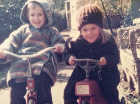 2 Kinder auf Fahrzeugen in Mänteln & Mützen, beide lachen.